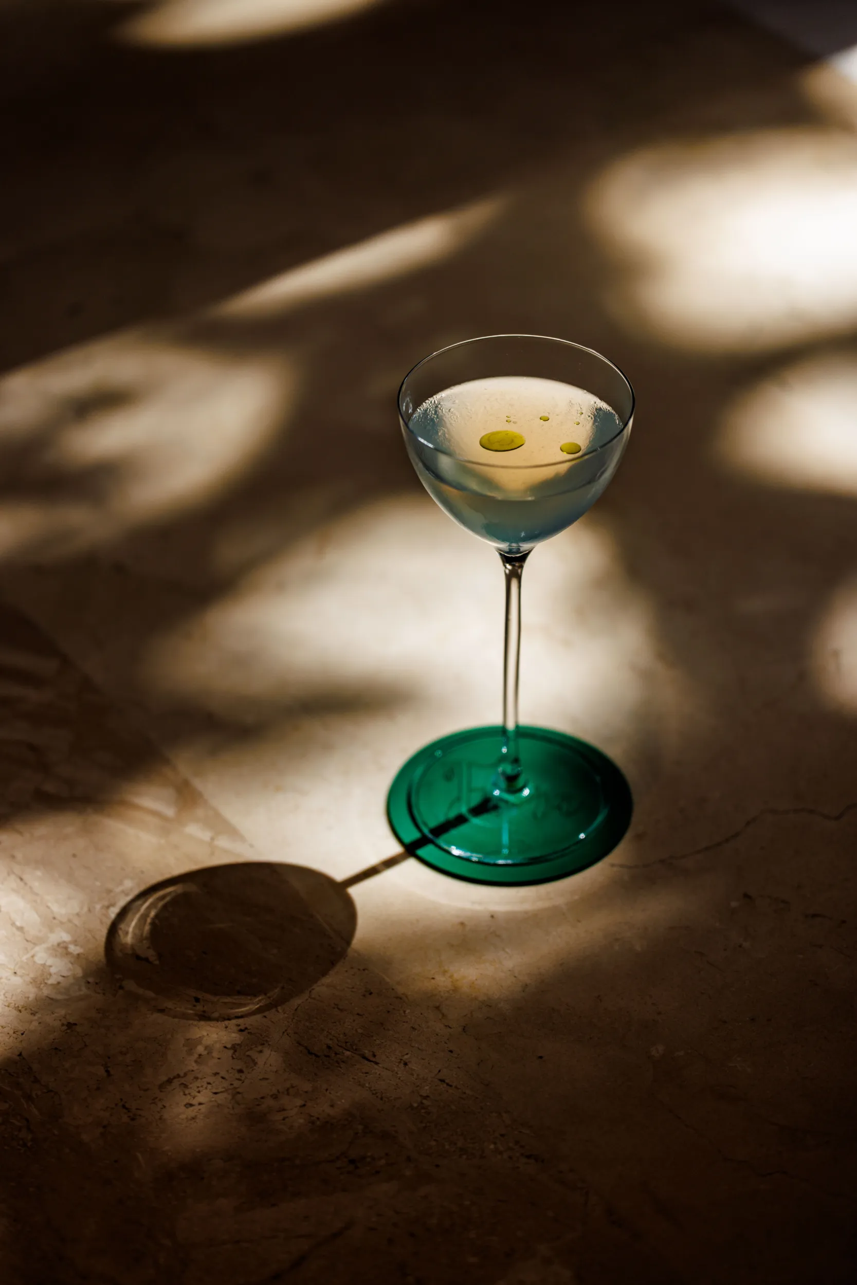 Green-tinted martini with an olive on a concrete floor in dappled sunlight
