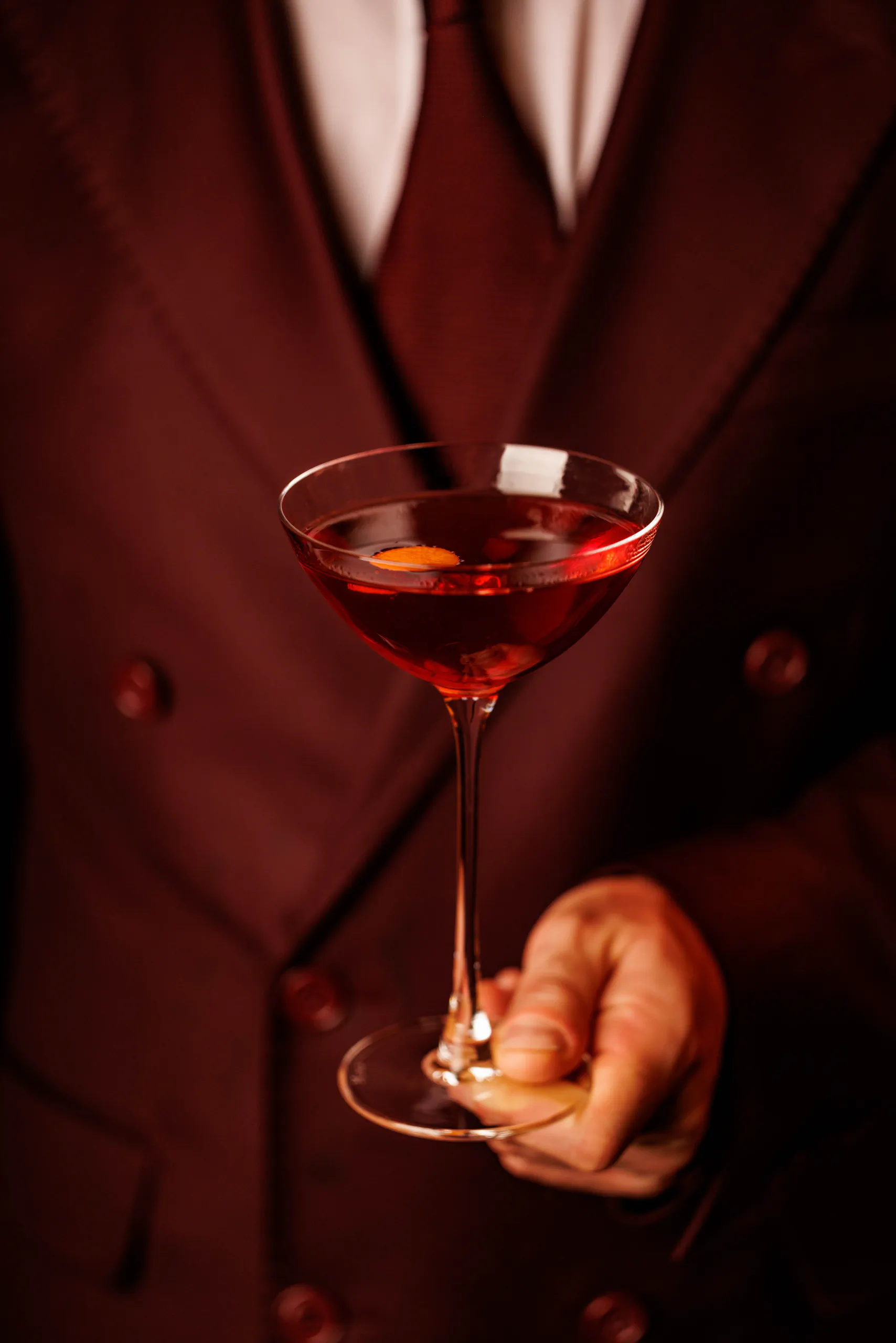 Person in a burgundy suit holding out a Manhattan cocktail in a coupe glass