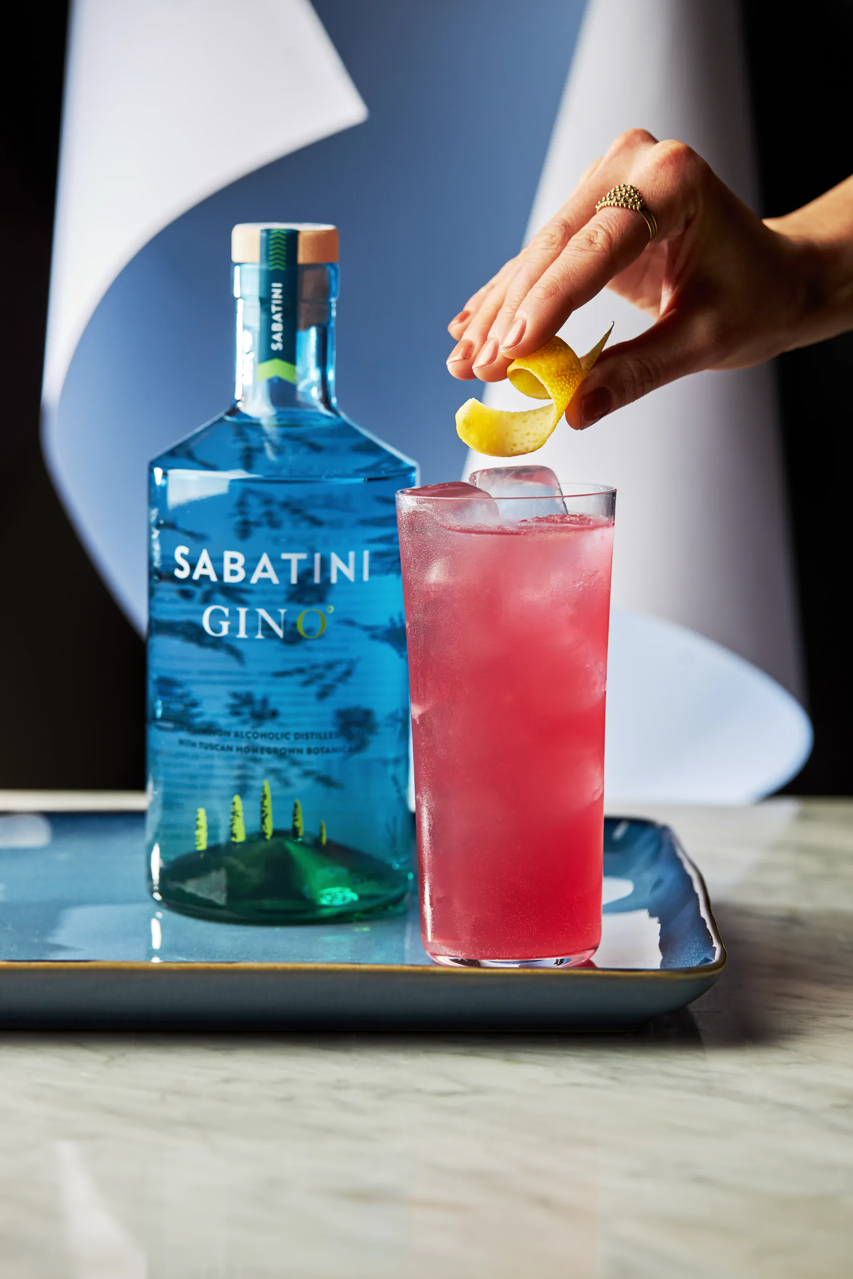 Hand squeezing lemon over a pink cocktail beside a bottle of Sabatini Gin