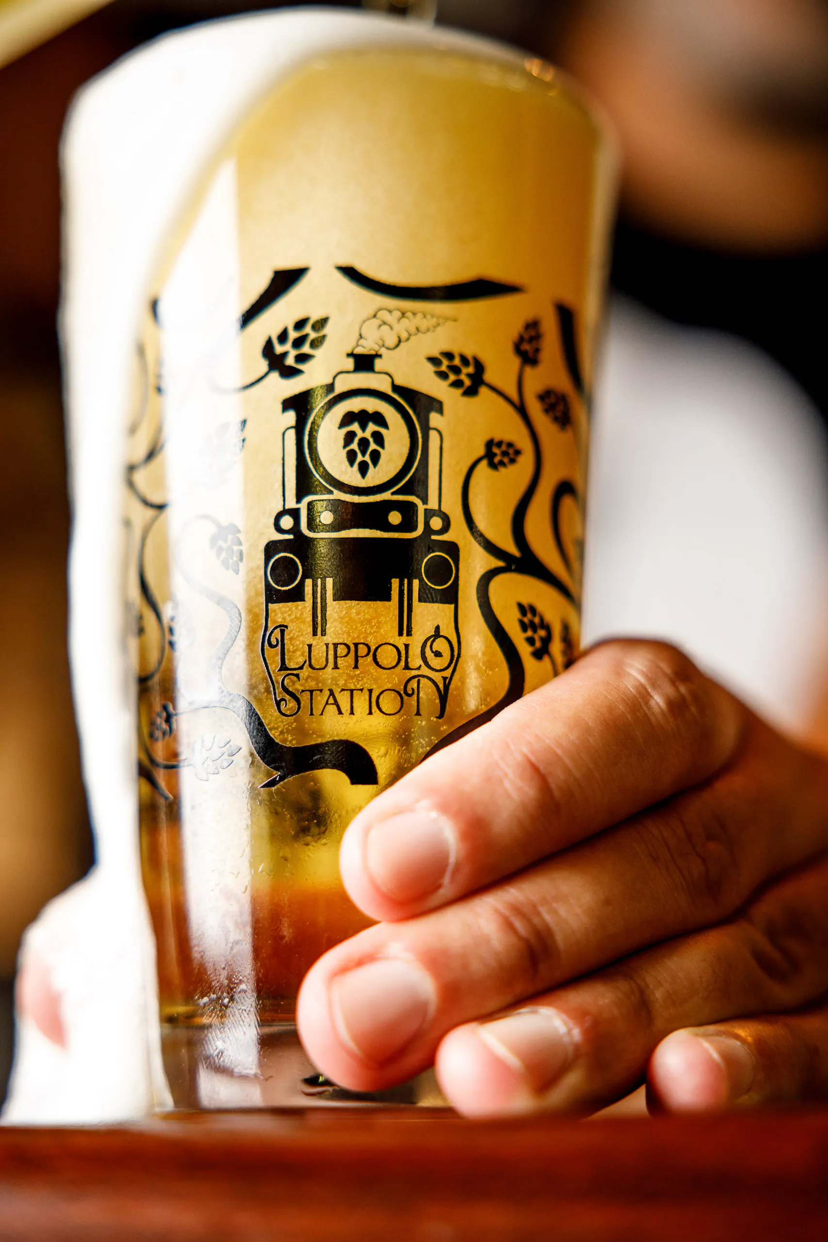 Hand holding a pint of golden beer with a printed Luppolo Station logo