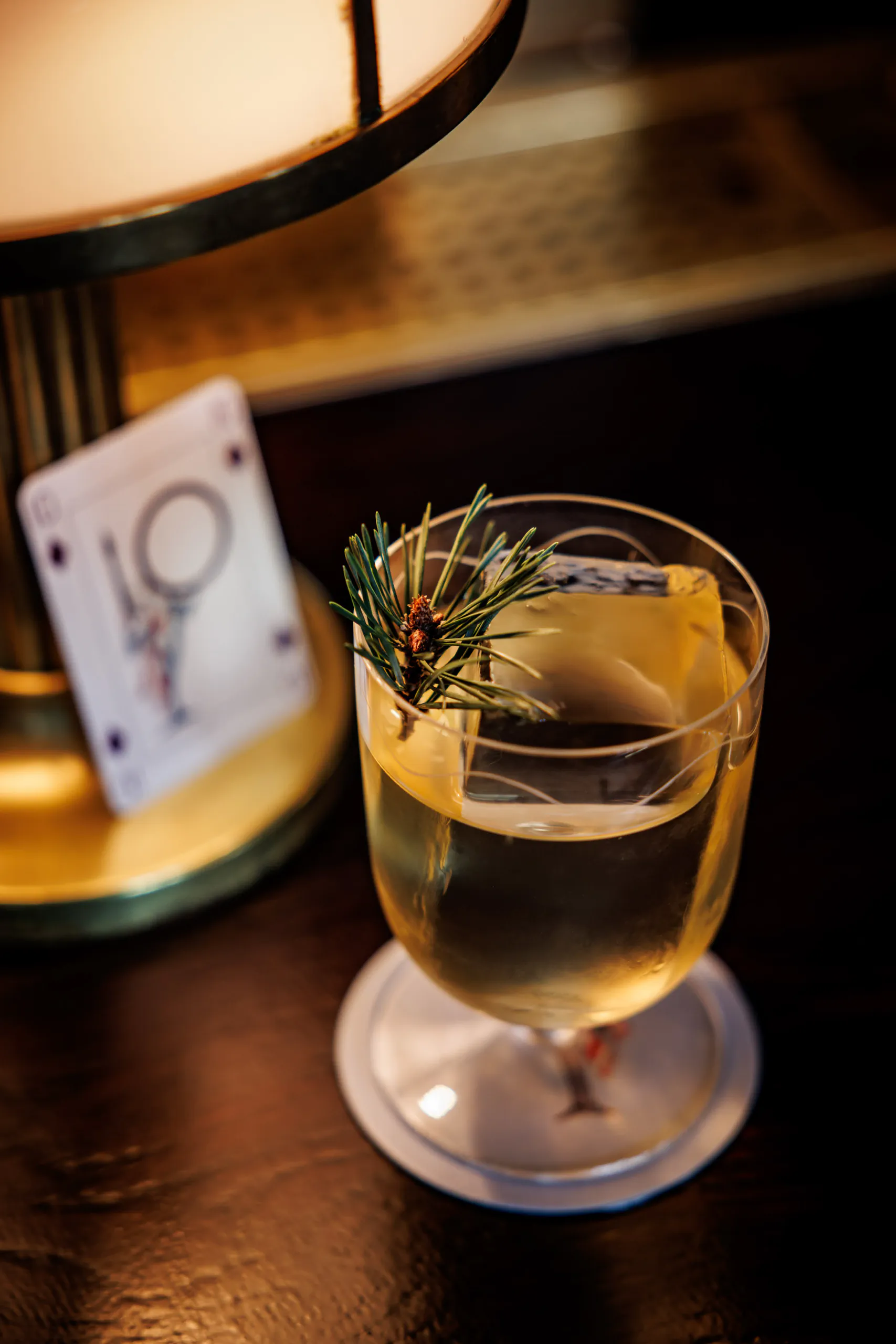 Rosemary-garnished cocktail in a stemmed glass beside a lamp and playing cards
