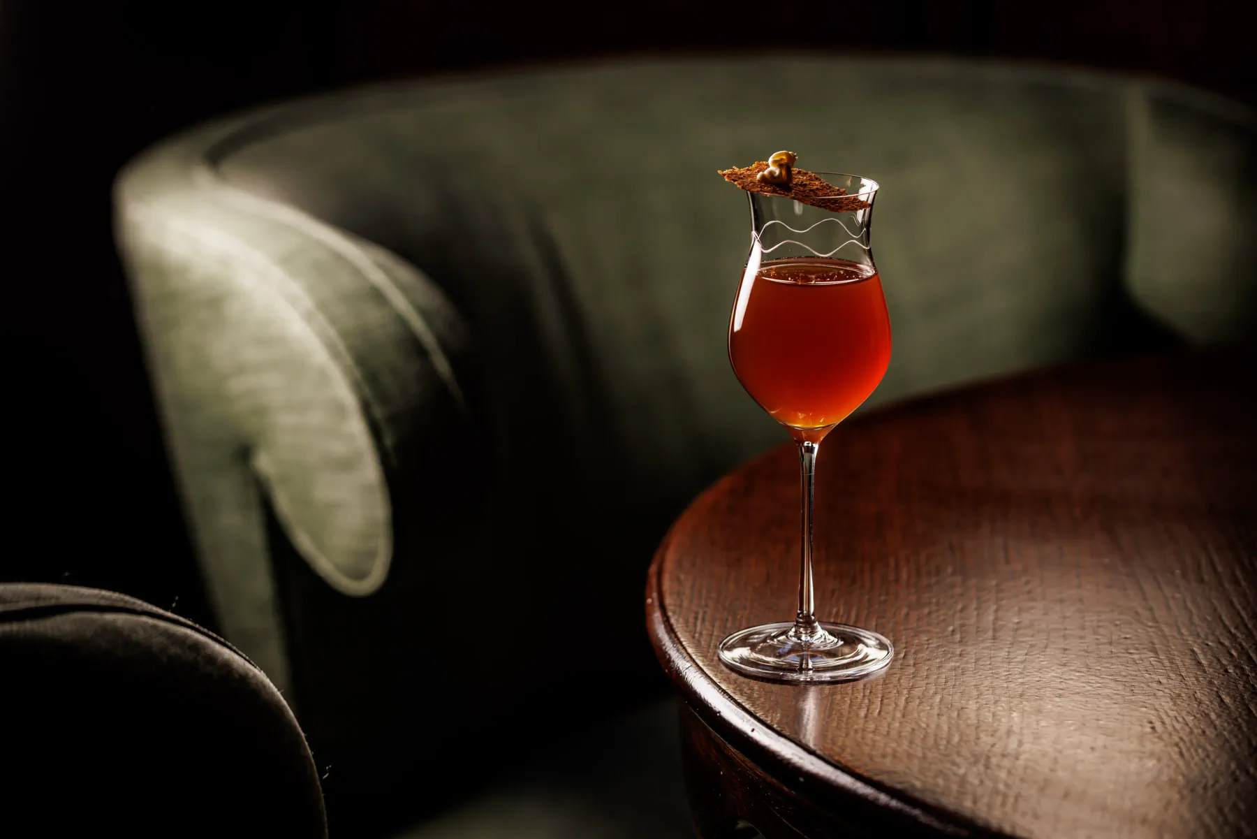 Amber cocktail in a stemmed glass on a side table beside a green velvet armchair at Scarfes bar in London, UK