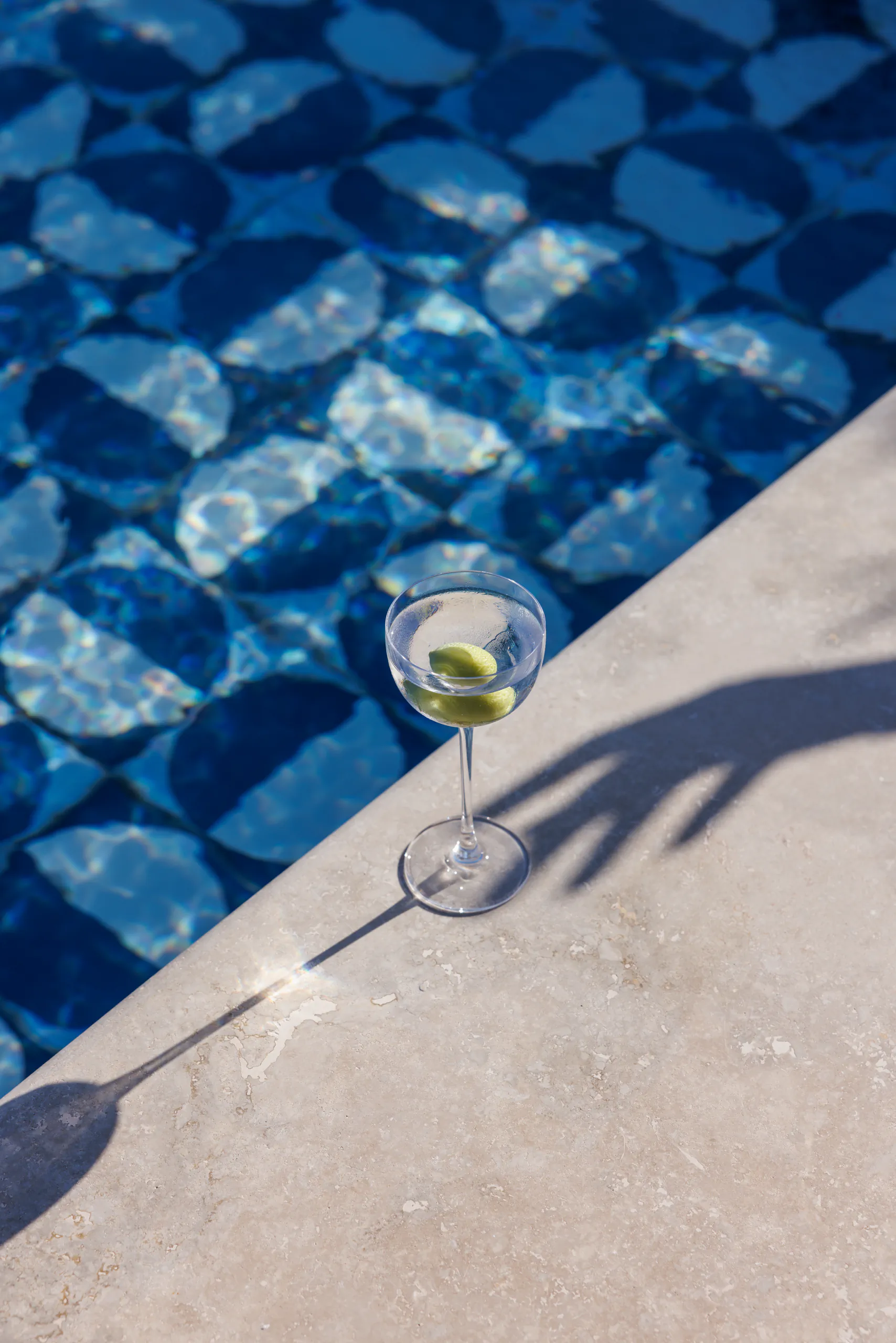 Martini with an olive at the edge of a blue-tiled swimming pool in sunlight