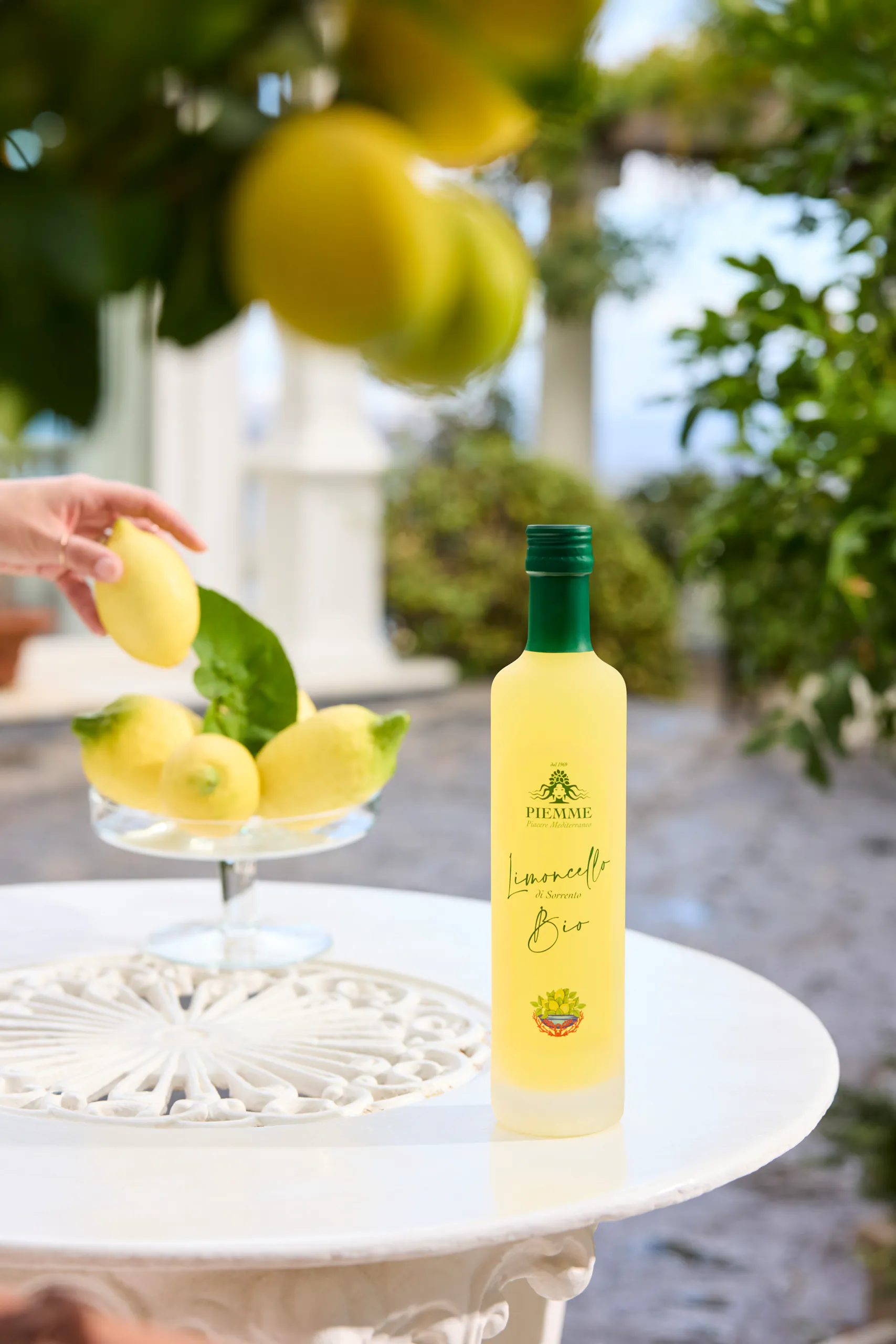 Hand arranging lemons beside a limoncello bottle under a lemon tree — Bellevue Syrene, Sorrento