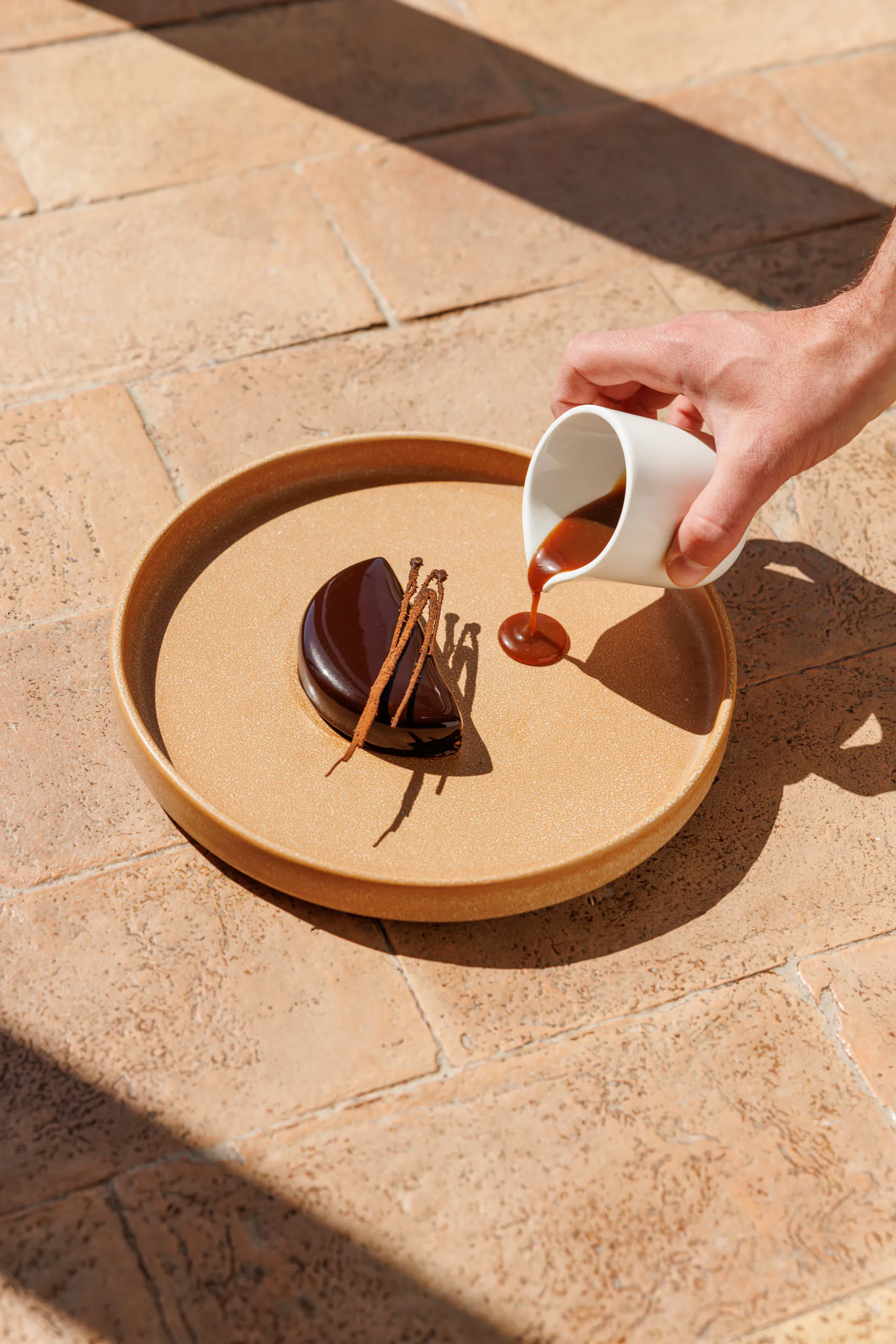 Hand pouring sauce over a chocolate dessert on a terracotta floor