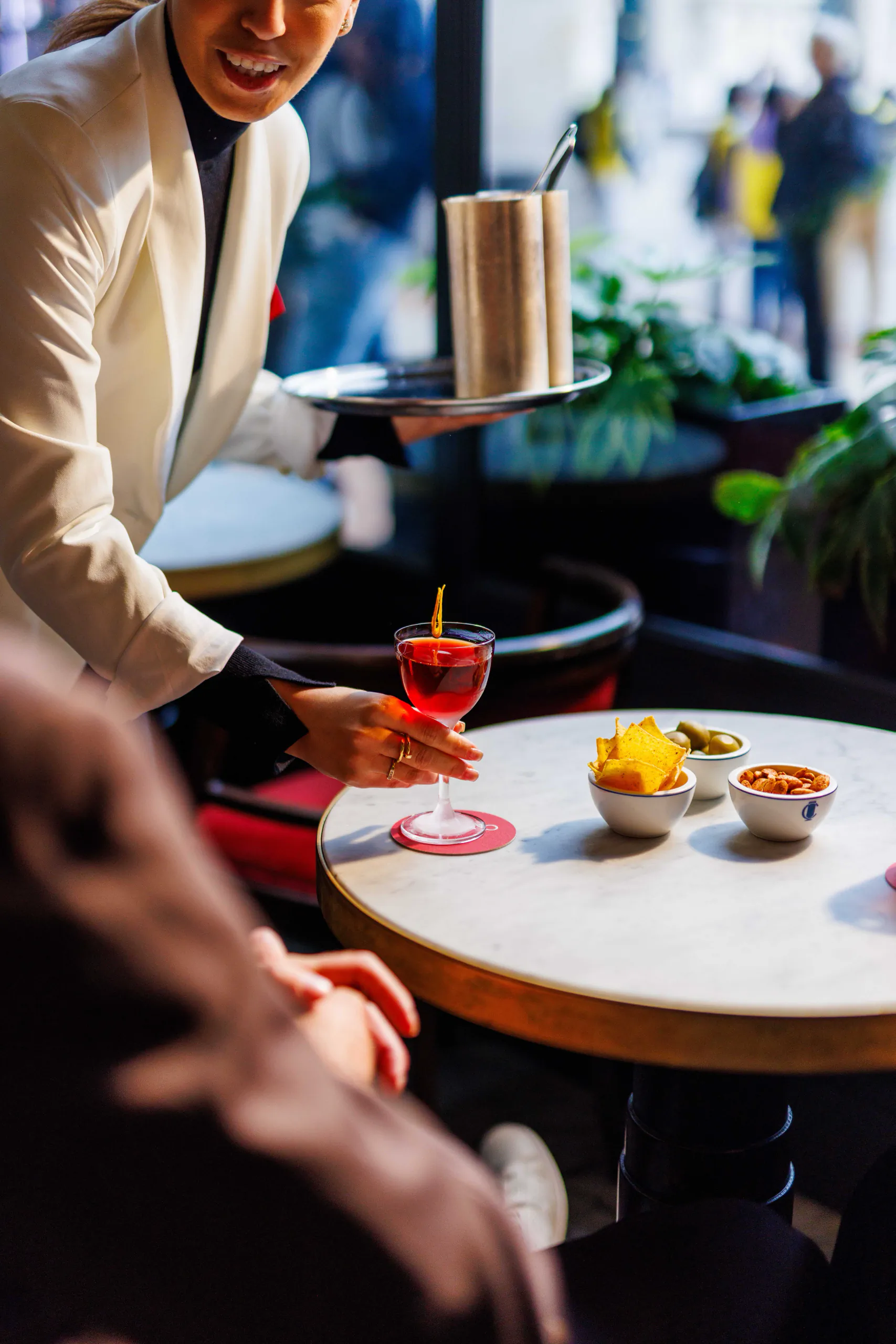 Server bringing a drink to a marble table with a red cocktail and snacks in a lively bar at Camparino in galleria, Milan, Italy