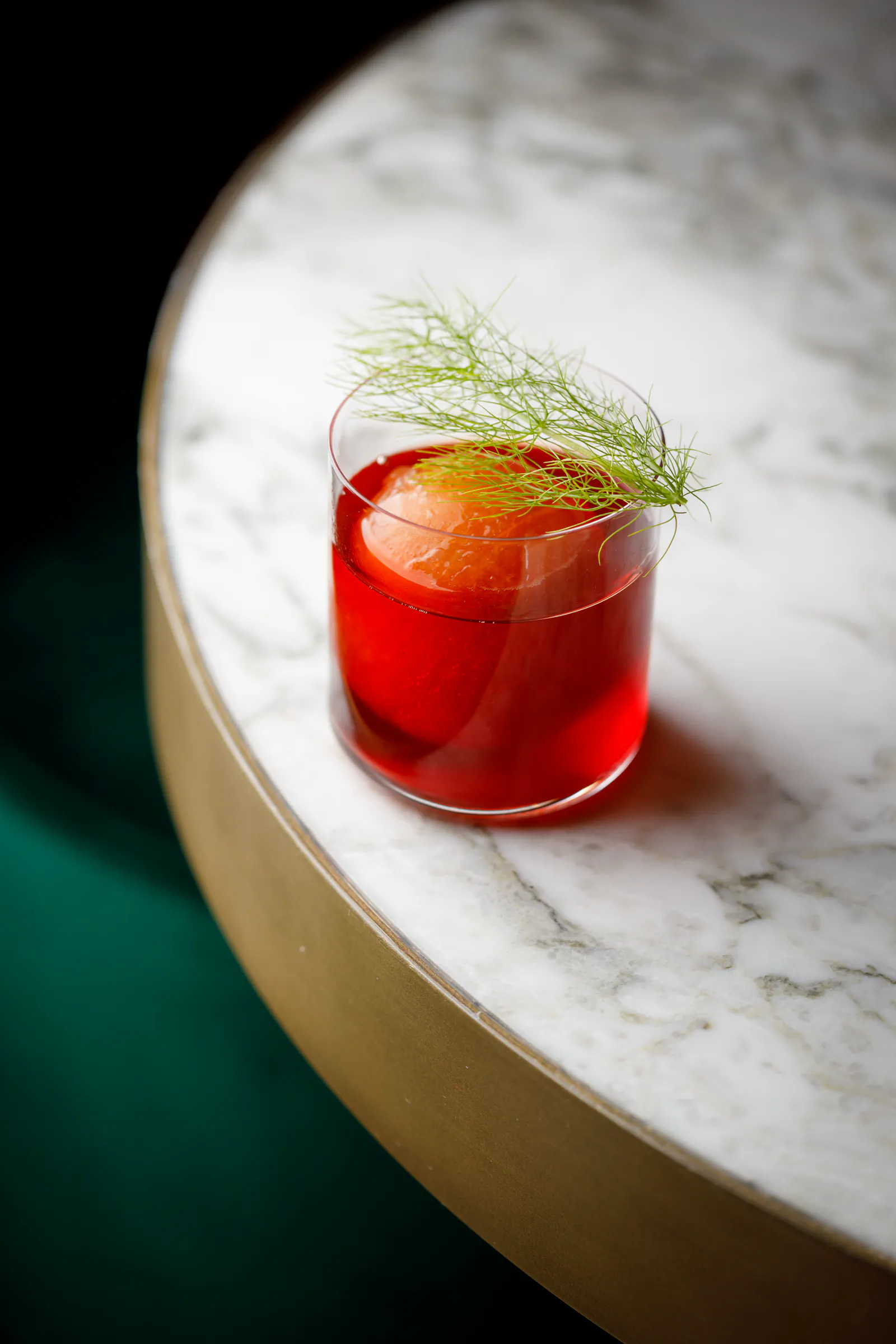 Red cocktail garnished with a fennel frond in a rocks glass on white marble at W Rome