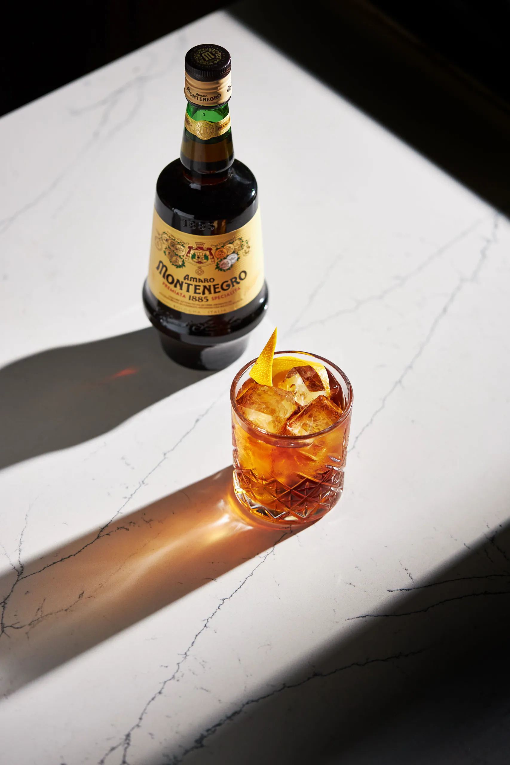 Bottle of Amaro Montenegro beside a glass of the liqueur over ice on sunlit marble