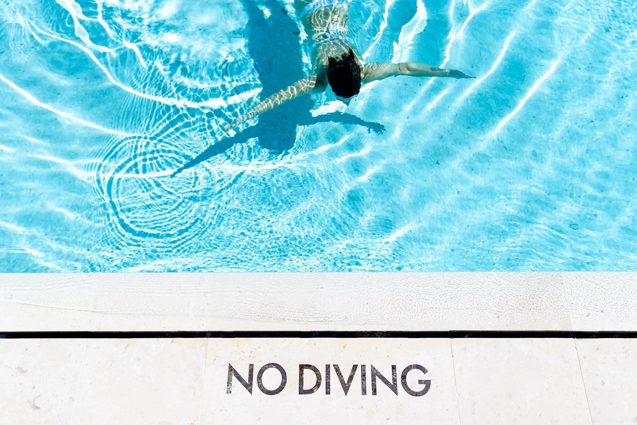 Person swimming in a turquoise pool seen from above, 'NO DIVING' on the deck