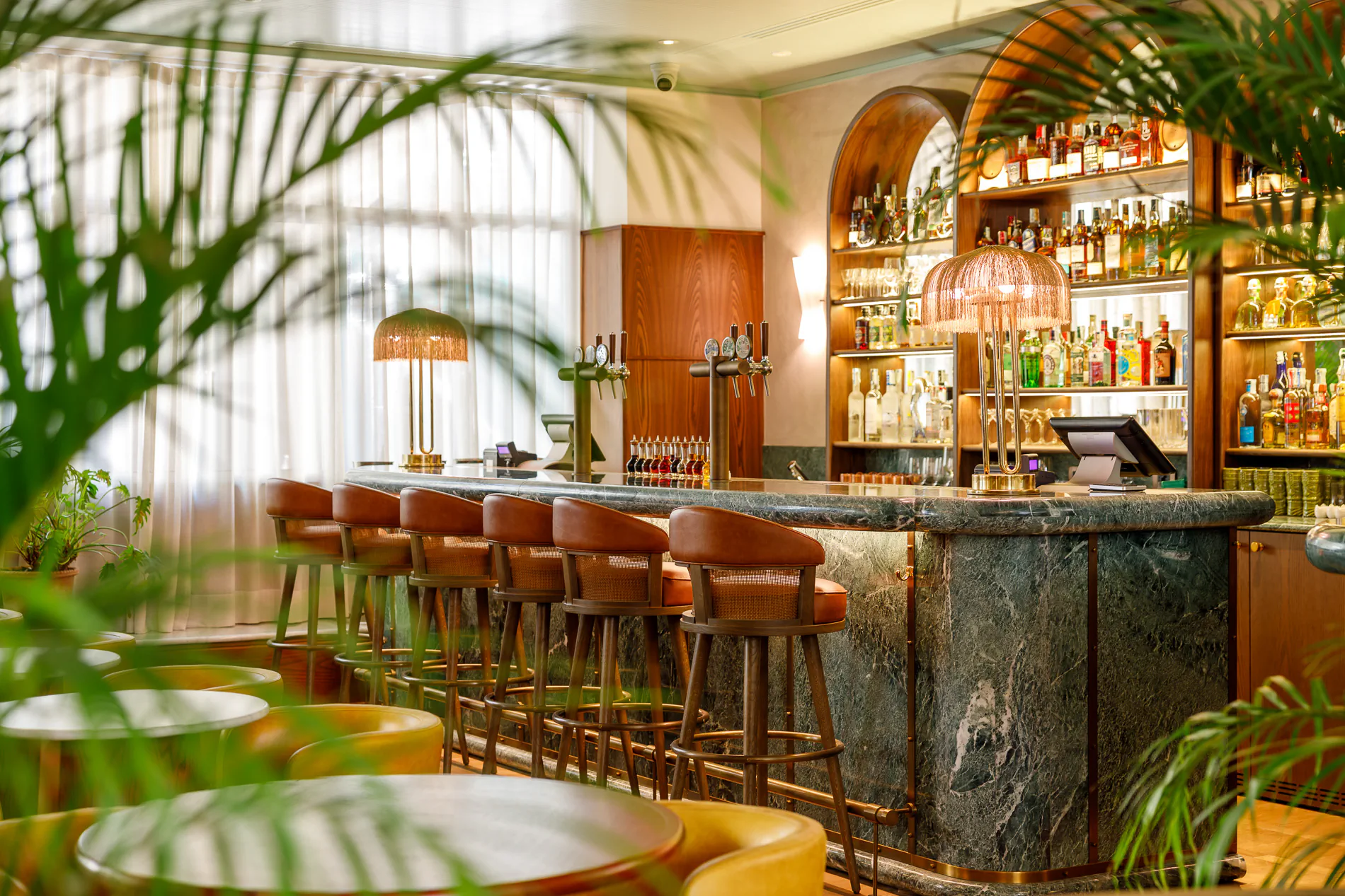 Lush bar with plants, a marble counter, high stools and shelves of spirits