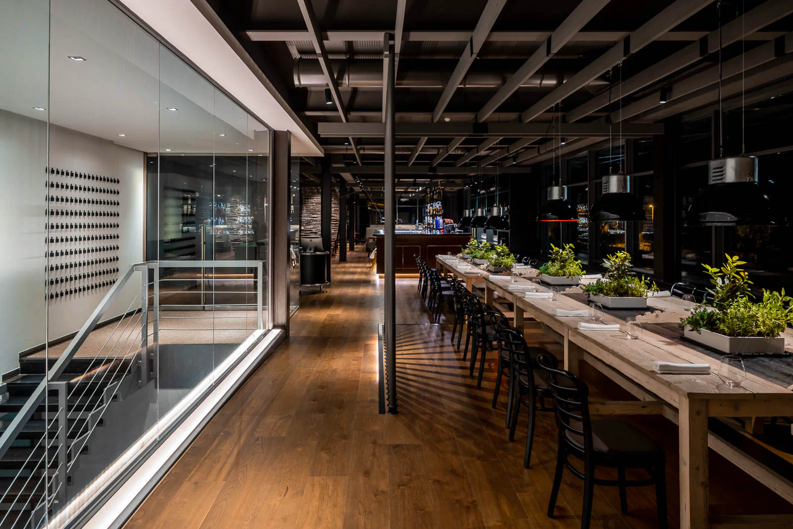 Long modern restaurant with a communal table lined with plants beside a glass wall