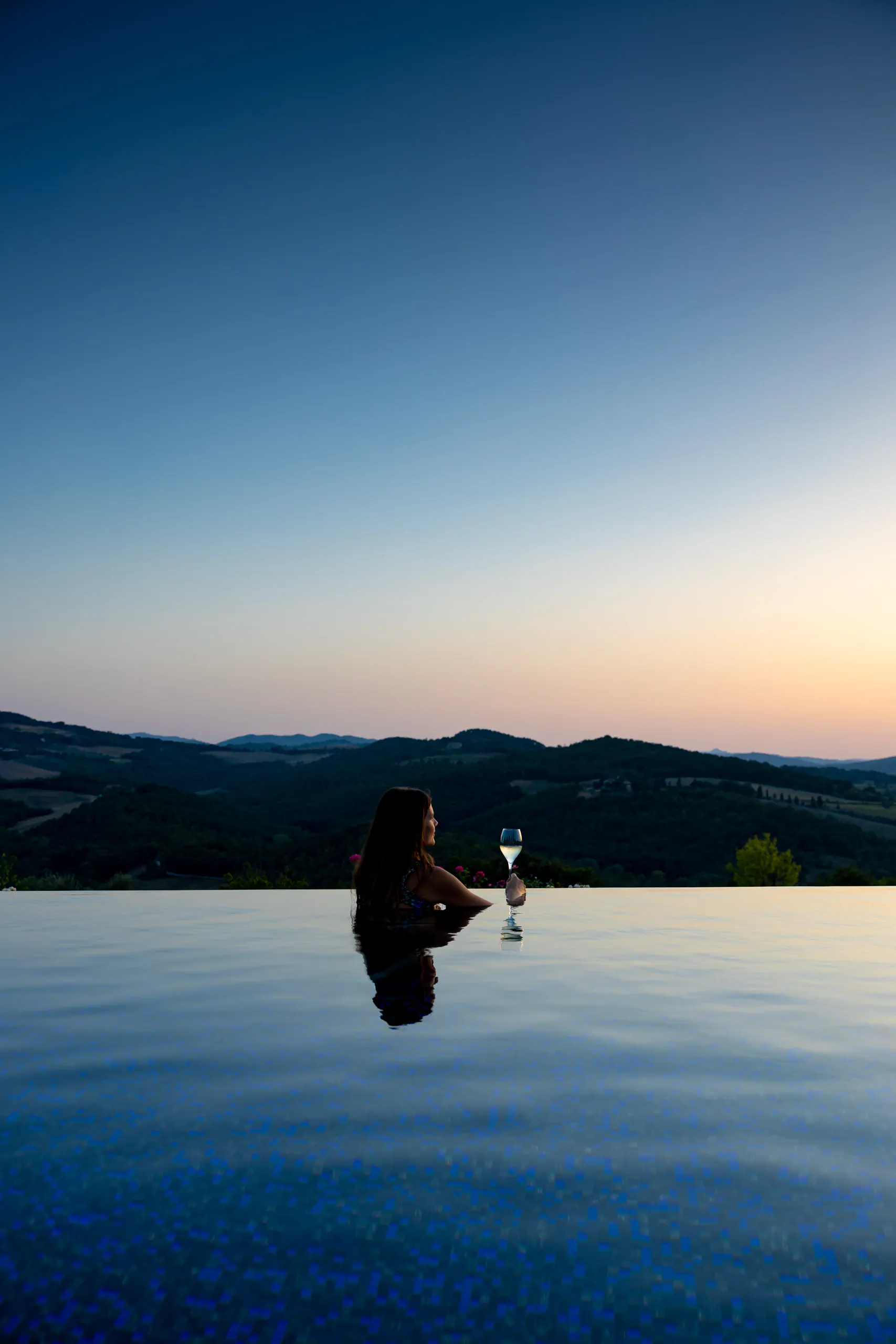 Person in an infinity pool at dusk holding a drink, overlooking the hills